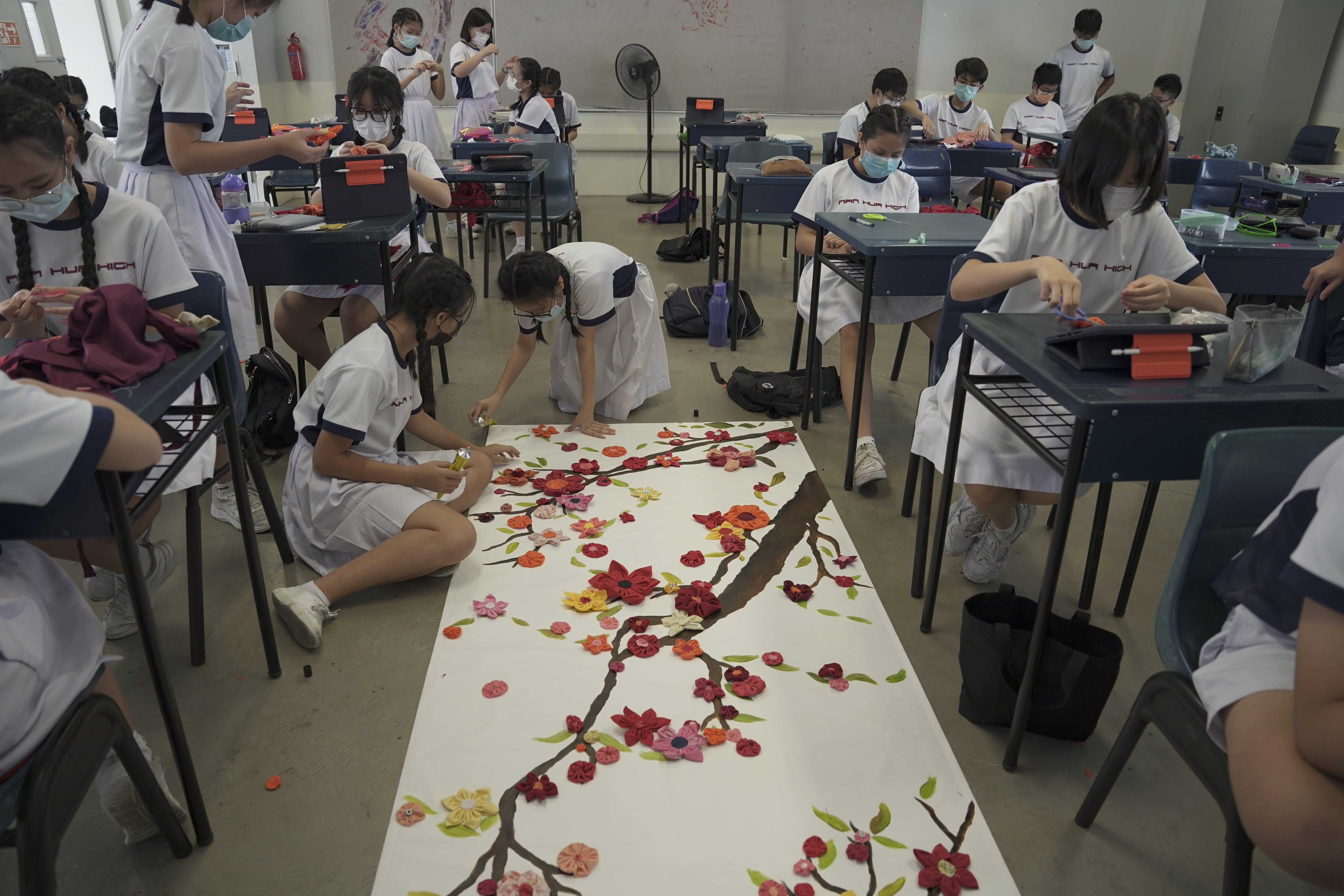 Students in white uniforms and masks arrange colorful fabric flowers on a large cherry blossom branch artwork on the floor.